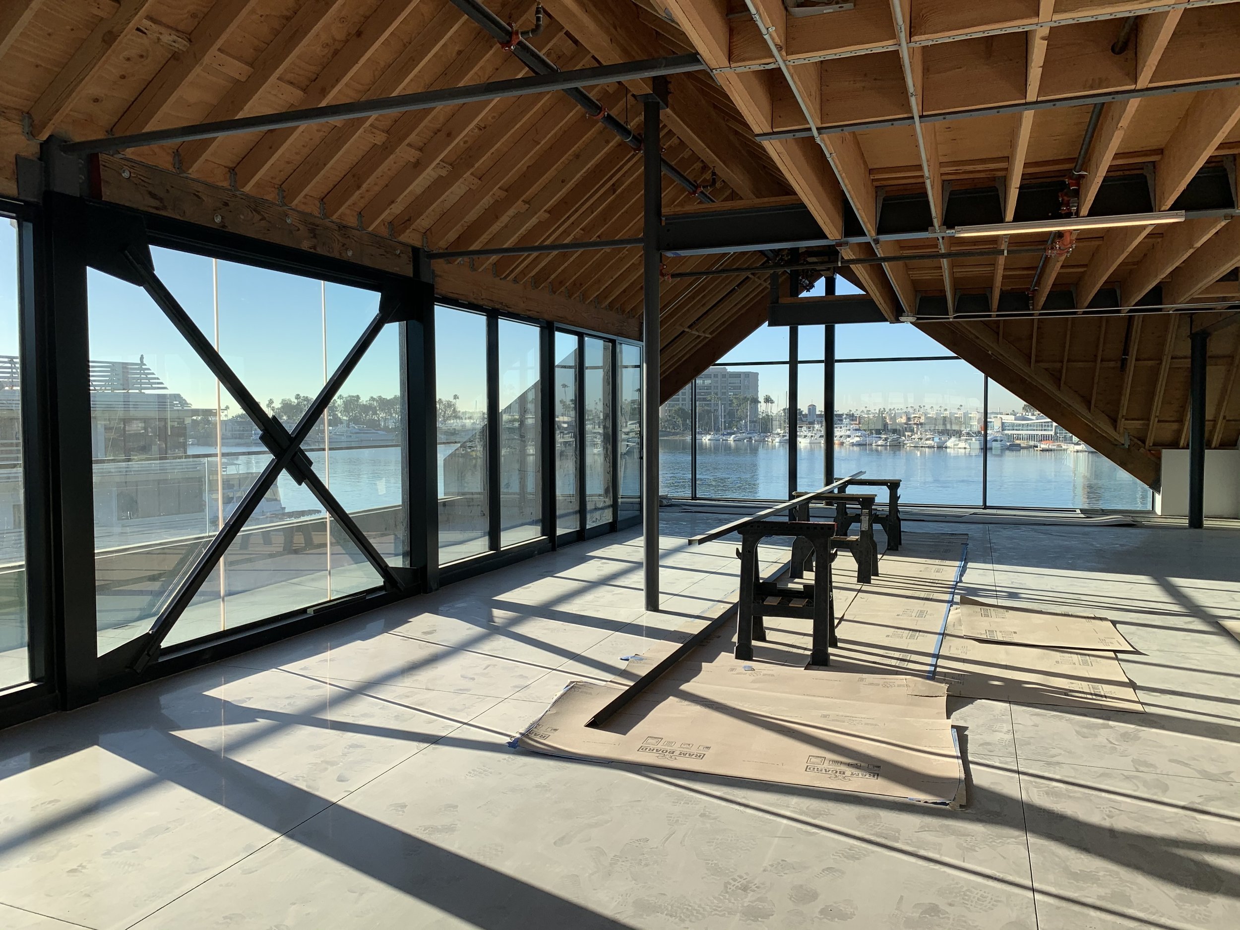 Interior of a modern building with large windows overlooking a marina, exposed wooden ceiling beams, and structural steel supports.
