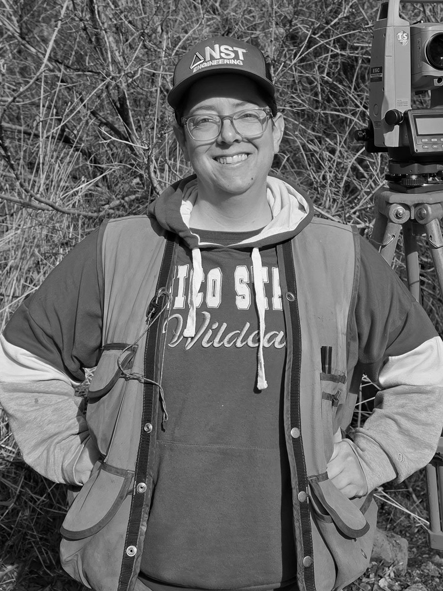 A person wearing a cap and outdoor work vest, standing next to surveying equipment.